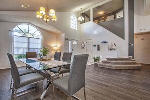 A spacious living area with a glass dining table, grey upholstered chairs, and a staircase leading to an upper level.