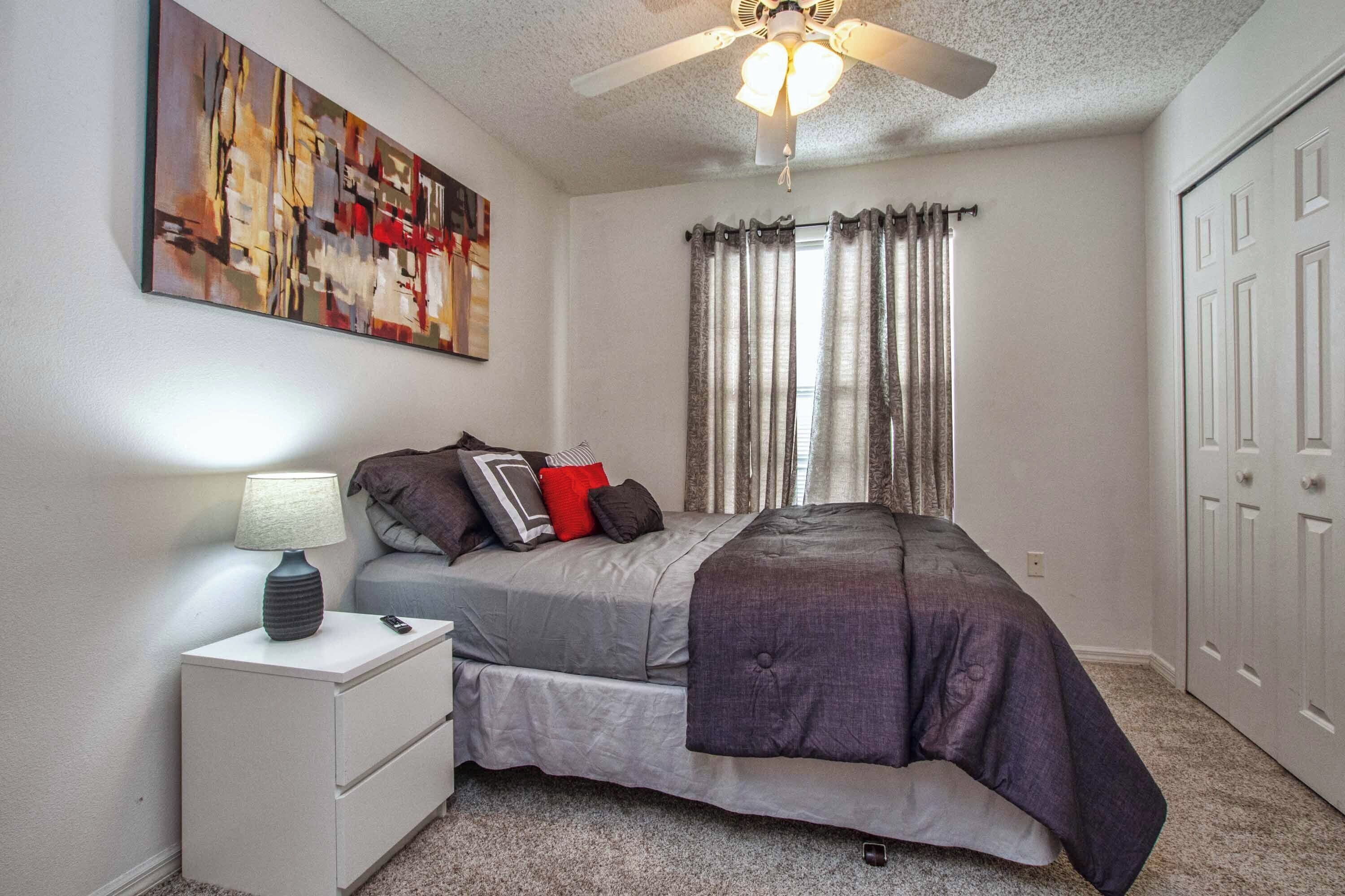 A bedroom with a bed, nightstand, lamp, ceiling fan, and a painting on the wall.