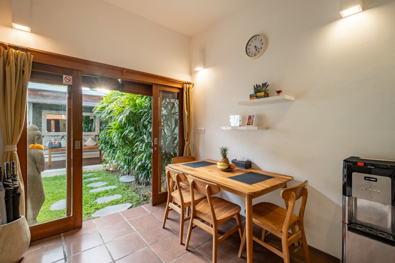 A small kitchen with a dining table, chairs, a refrigerator, and a clock on the wall.