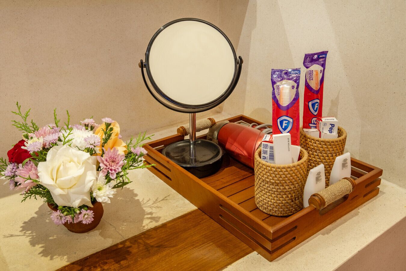 Una bandeja de madera con un espejo redondo, un jarrón de flores y dos cestas con artículos de aseo personal.