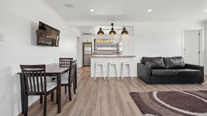 Una cocina moderna con armarios blancos, un refrigerador de acero inoxidable y una barra con taburetes. Hay una mesa de comedor con sillas y un sofá de cuero negro en la sala de estar.