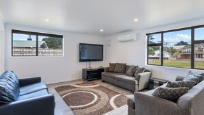 Una sala de estar moderna con televisor de pantalla plana, dos sofás, una mesa de centro y un ventanal con vistas a casas y zonas verdes.