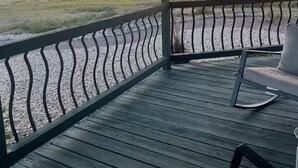 Una terraza de madera con barandilla metálica, con vista a un campo cubierto de hierba y un atardecer de fondo.