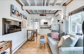 Eine moderne Küche mit weißen Schränken, ein Essbereich mit Holztisch und -stühlen sowie ein Wohnzimmer mit grauem Sofa und Kissen.