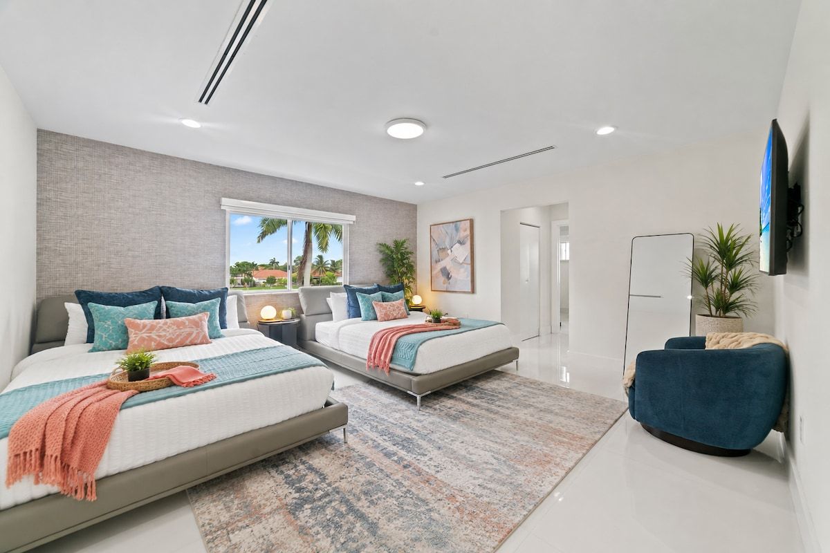 A modern bedroom with two beds, a large window, and a flat-screen TV mounted on the wall.