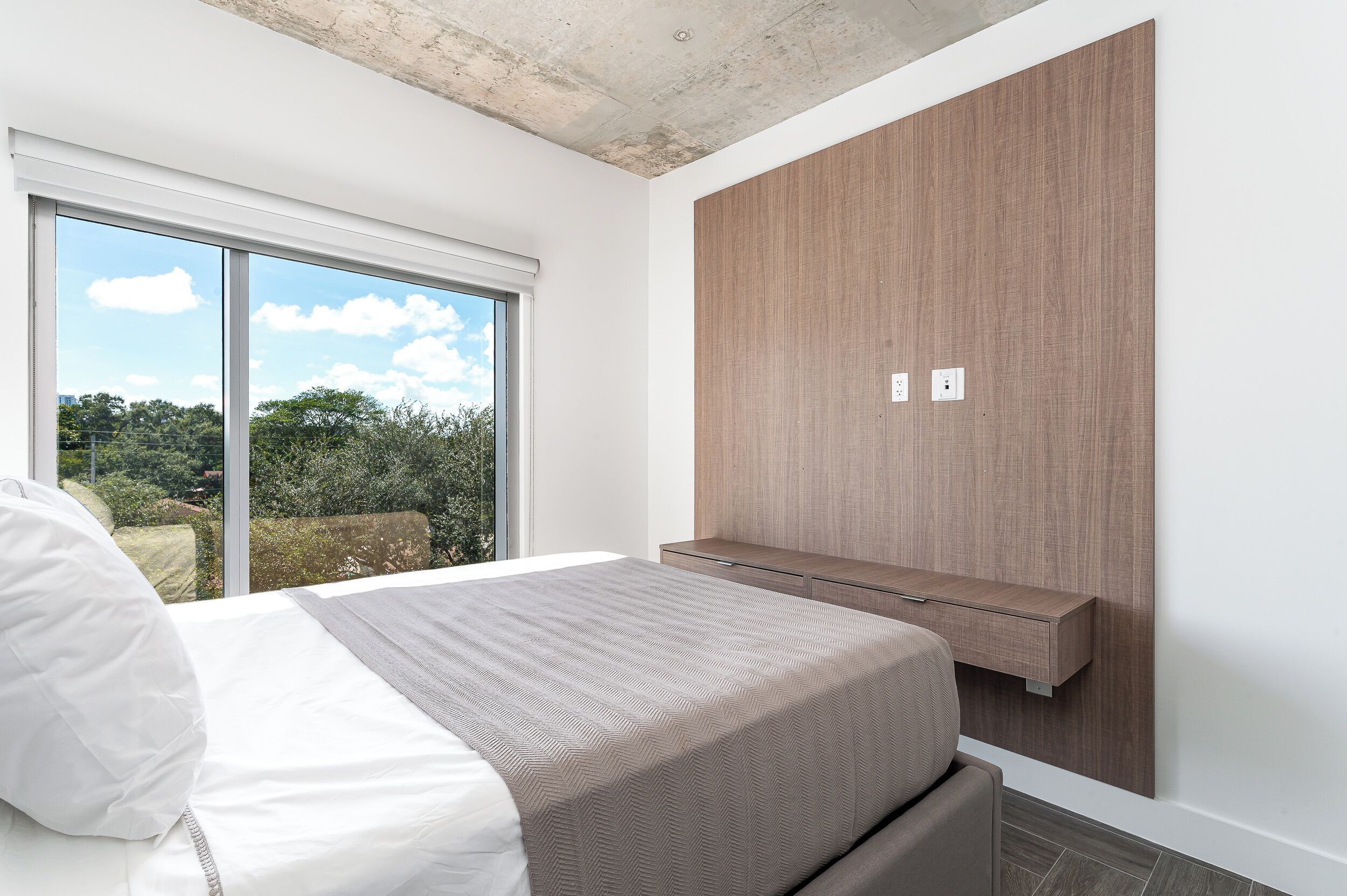 A bedroom with a large window, a bed with white bedding, a wooden wardrobe, and a small bedside table.