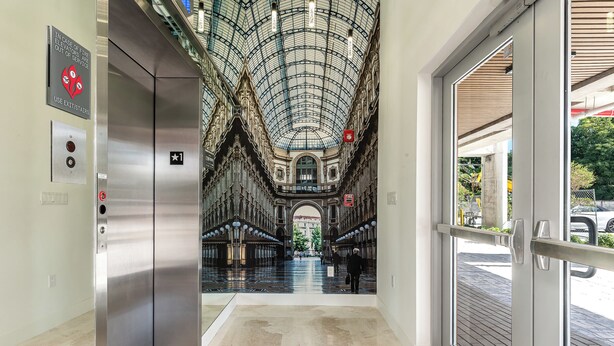 An interior corridor with a glass ceiling, an elevator, and a door leading outside. - Milan Aventura Hotel (Ojus)