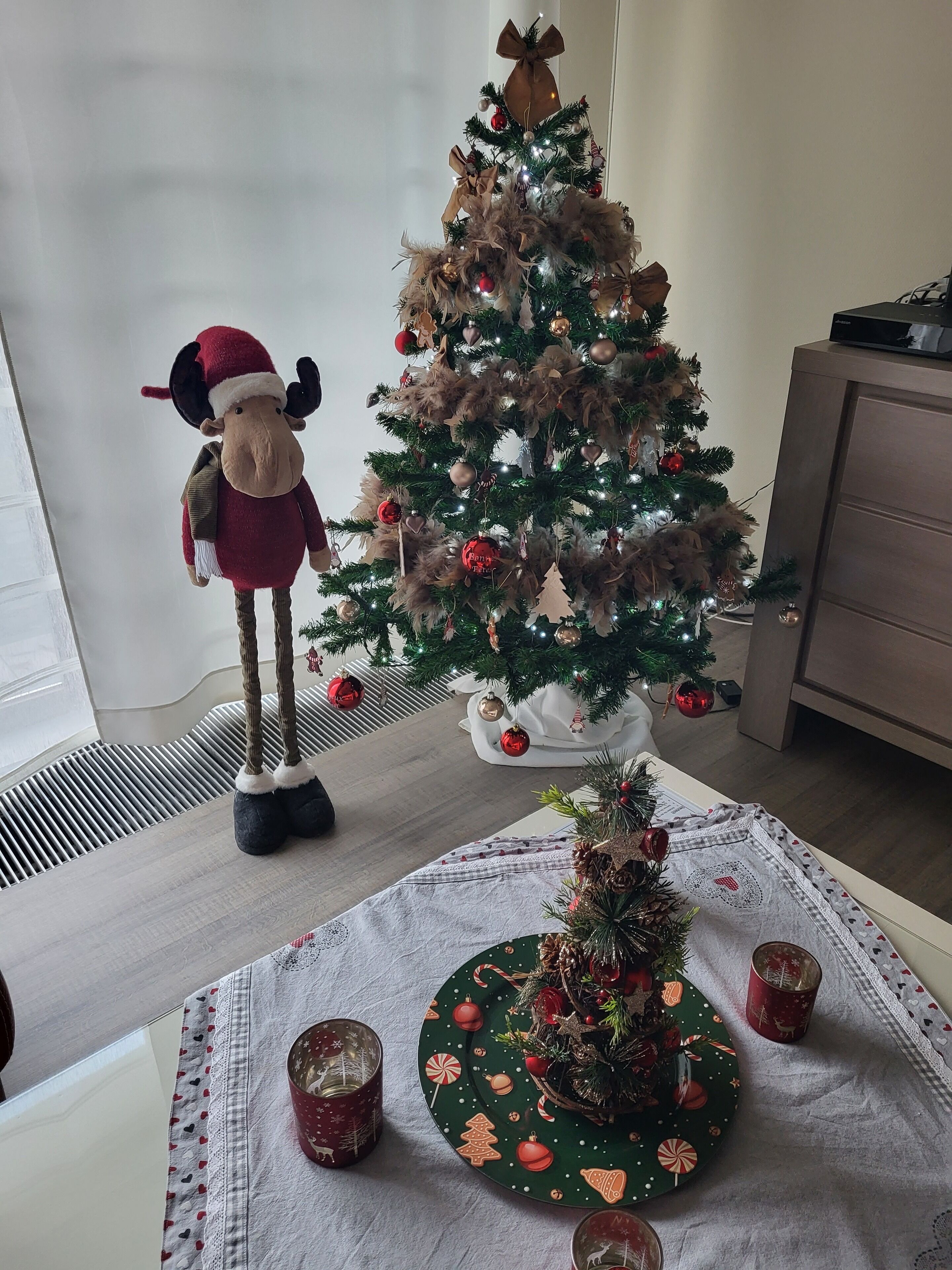 Een kerstboom met versieringen en lichtjes, een pluche speelgoed en een tafel met een placemat en kaarsen.