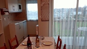 Een eetgedeelte met een tafel gedekt voor twee personen, een keuken op de achtergrond en een uitzicht op een woonwijk door een raam.