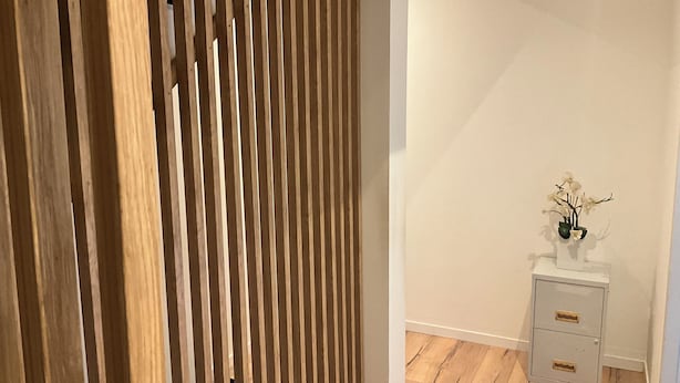 A room with a wooden slat partition, a white wall, a small white cabinet with drawers, and a vase with flowers on top.