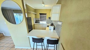 Une cuisine de taille réduite avec un plan de travail blanc, deux chaises noires et un sol carrelé.