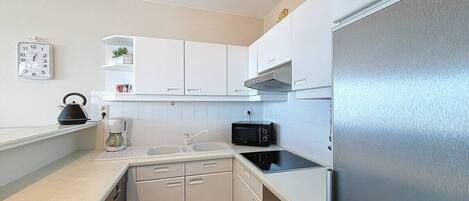 Una cocina compacta con armarios blancos, un refrigerador de acero inoxidable, un microondas y una tetera sobre la encimera.