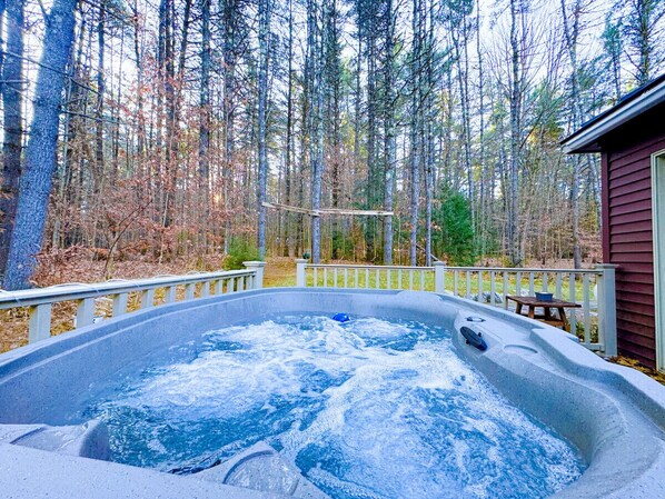 A hot tub surrounded by a wooden deck with a railing, set against a backdrop of trees and a building. - Luxe Lodge in the Woods! With Sauna & Hot tub (Conway)