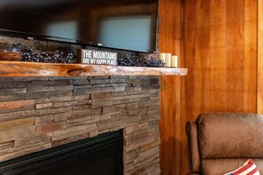A stone fireplace with a sign above it, a television mounted above the fireplace, and a wooden wall in the background. - Luxe Lodge in the Woods! With Sauna & Hot tub (Conway)