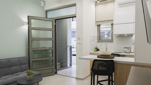 A modern kitchen with a built-in stove, a sink, and a window above the counter. There is a dining area with a grey sofa and a small table with a plant. - Peris #5 w. Yard steps to Bus St. & Old Town (Chania, Crete)