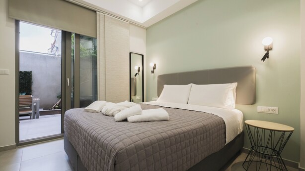 A bedroom with a bed, a round side table, a sliding glass door leading to a balcony, and a wall-mounted light fixture. - Peris #5 w. Yard steps to Bus St. & Old Town (Chania, Crete)