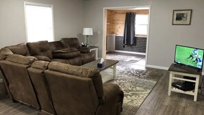 Una sala de estar con un sofá seccional marrón, una mesita con una lámpara, un televisor sobre un mueble bajo y vista a un interior de madera a través de un umbral abierto.