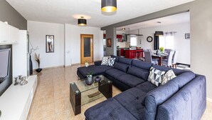 Een moderne woonkamer met een hoekbank, een glazen salontafel en een televisie. Achtergrond is een keuken met rode kastjes en een eethoek met een tafel en stoelen.