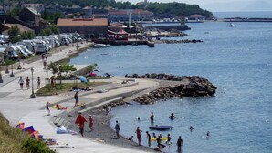 Ett område vid kusten med en asfalterad gångväg, en strand och möjlighet till aktiviteter på vattnet. Det står husbilar parkerade längs sidan, och en skylt som förbjuder hundar är synlig.