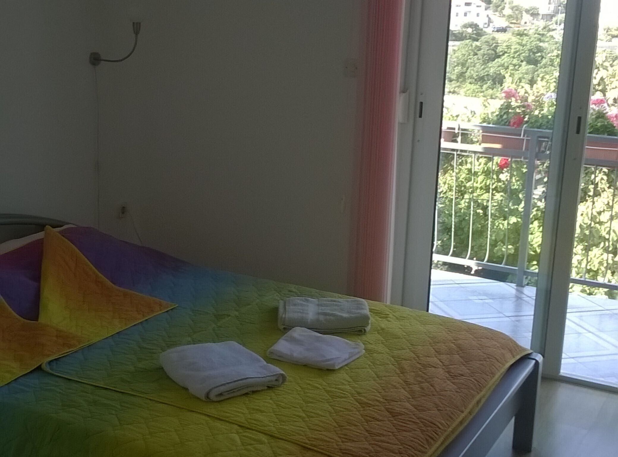 A room with a bed, a window, and a view of a balcony with plants.