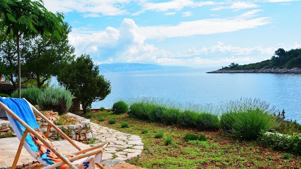 Un sentier de pierres mène à une plage où se trouvent une chaise et une serviette, d’où l’on peut admirer une mer calme et des montagnes lointaines.