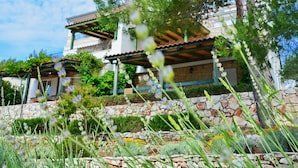 Une maison en pierre à deux étages, avec des fondations en pierre, un toit recouvert de tuiles et un jardin où poussent diverses plantes et fleurs.