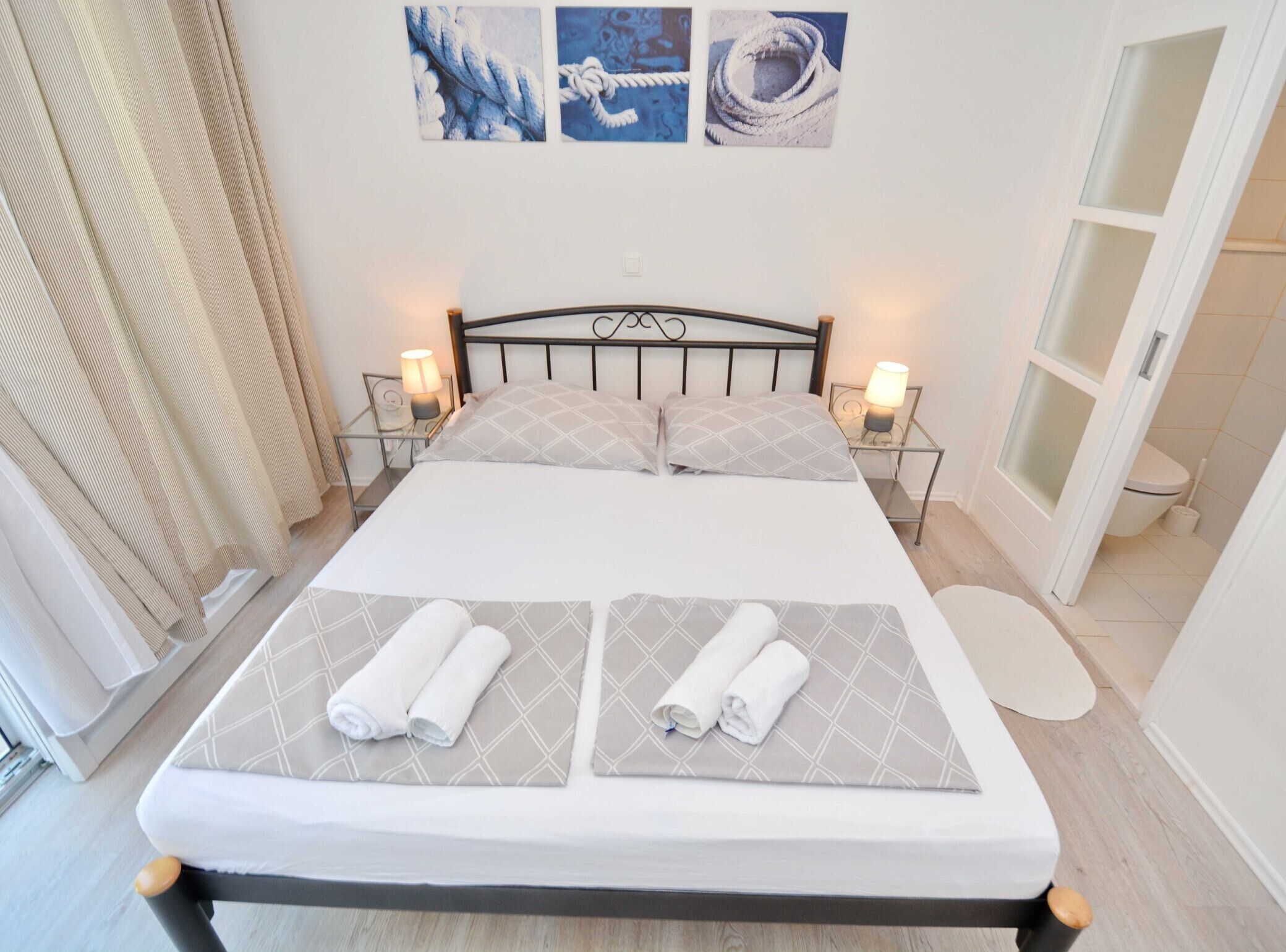 A neatly made bed with two pillows and two rolled towels, a bedside table with a lamp, and a wall with three framed pictures.
