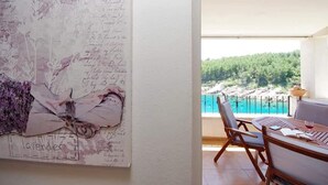 A room with a large floral artwork on the wall, a balcony with a view of a forest and sea, and a table with chairs.