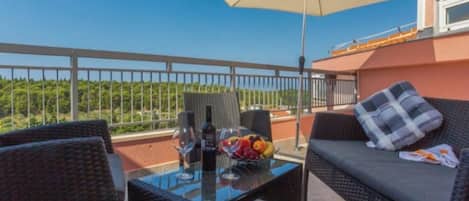 A balcony with wicker furniture, a glass table, a bottle of wine, and a bowl of fruit.
