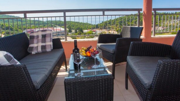 Un balcon agrémenté de meubles en osier, une table en verre avec une bouteille et des fruits, et une vue sur des collines verdoyantes.