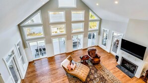 A spacious living room with a high ceiling, large windows, a fireplace, and a television.