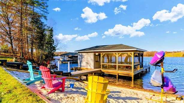 A lakeside deck with colorful Adirondack chairs, a dock, and a wooden structure with a glass enclosure.