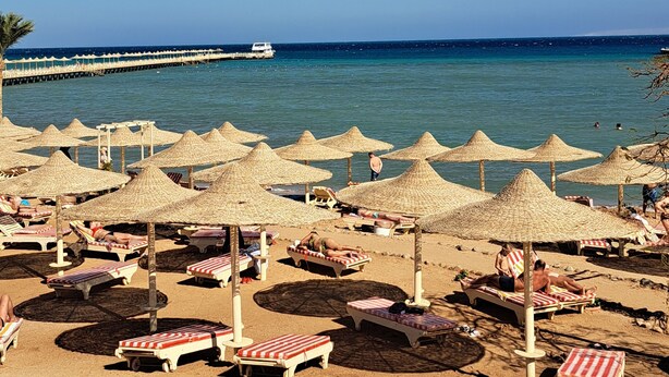 A beach with numerous sun umbrellas and lounge chairs, a pier extending into the sea, and clear blue skies.