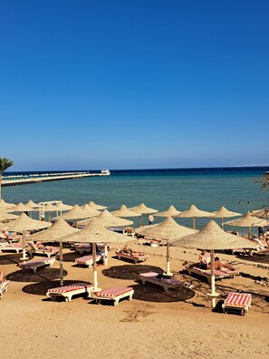 Una platja amb nombroses parasols i gandules, un moll que s'endinsxa al mar, i cels blaus i nets.