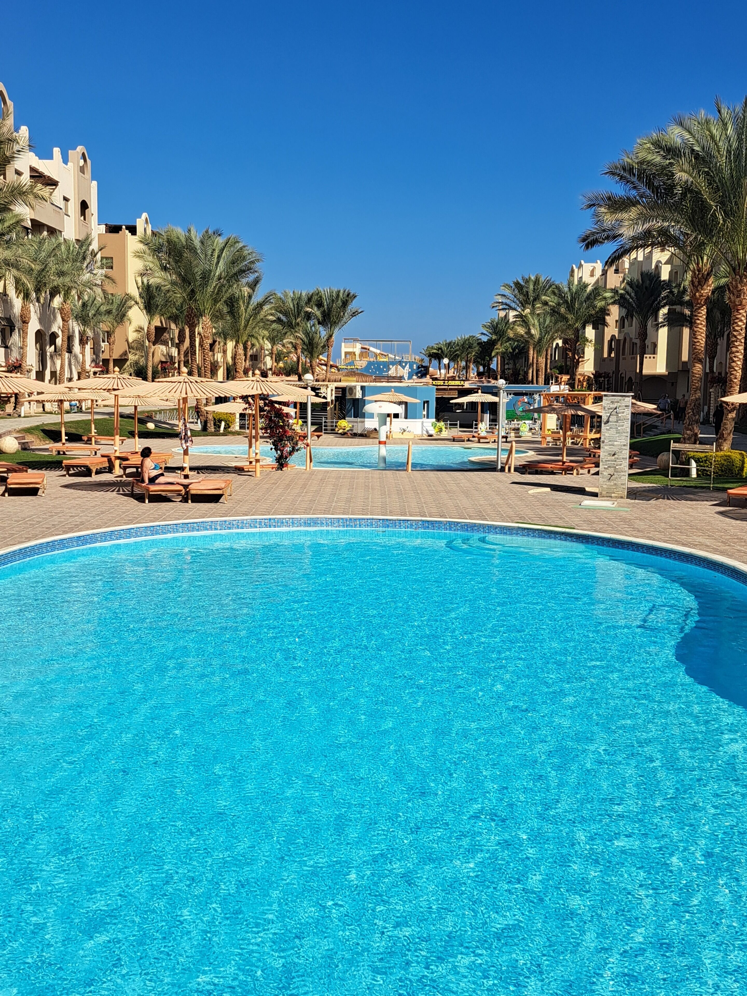 A large swimming pool with lounge chairs and umbrellas, surrounded by palm trees and a building with arched windows.