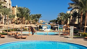 A large swimming pool with lounge chairs and umbrellas, surrounded by palm trees and a building with arched windows.