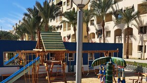 A playground with slides and climbing structures, surrounded by palm trees and a building with balconies.