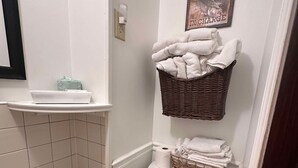 Un baño con inodoro, lavamanos, cubo de basura y una cesta con toallas en la pared.