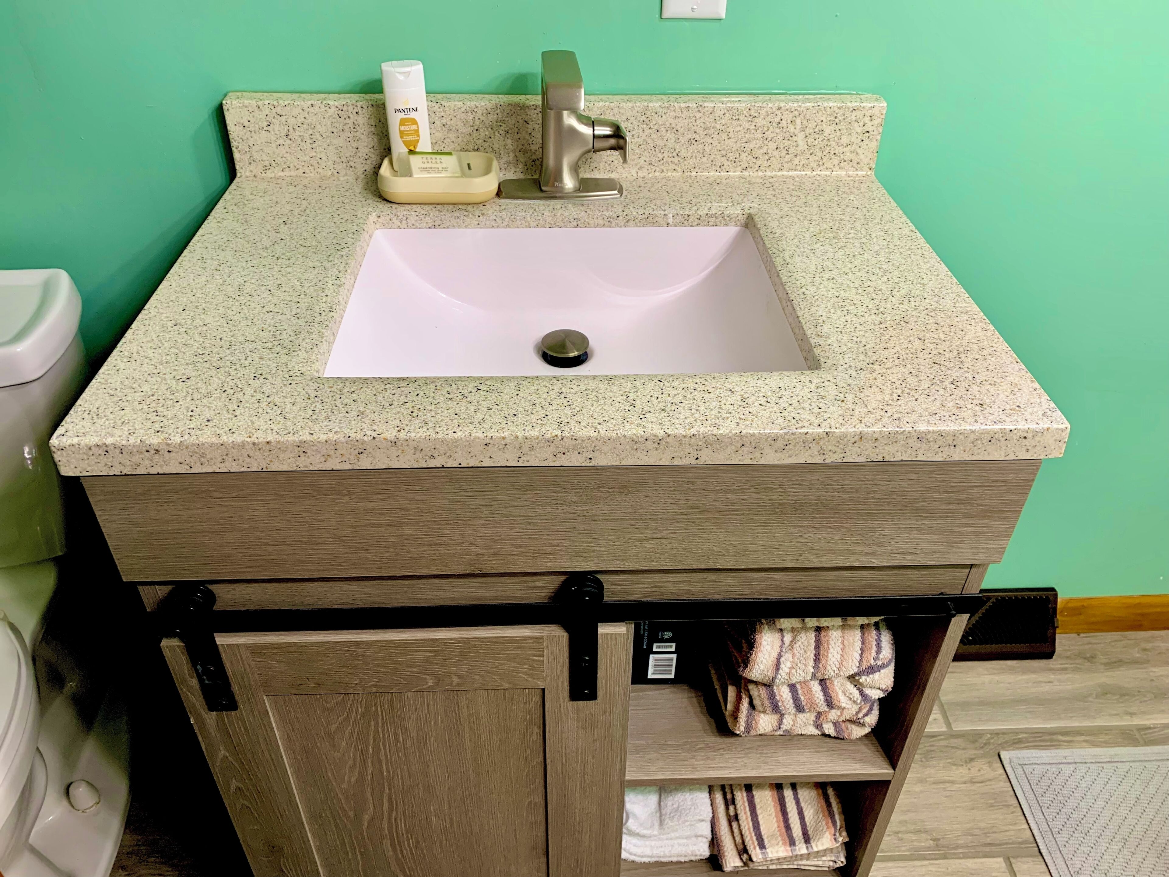 A bathroom with a beige granite countertop, a white sink, a faucet, and a toilet. There is a towel rack with folded towels and a bottle of Pantene shampoo.