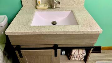 A bathroom with a beige granite countertop, a white sink, a faucet, and a toilet. There is a towel rack with folded towels and a bottle of Pantene shampoo.