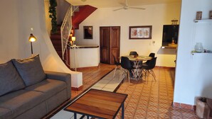 A living room with a sofa, a coffee table, a staircase, a dining area with a table and chairs, and a kitchen area with a refrigerator and cabinets.