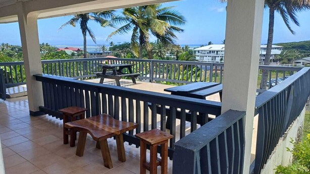 A covered outdoor area with wooden tables and chairs, a railing, and a view of the ocean and palm trees. - Bula Retreat (Sigatoka)