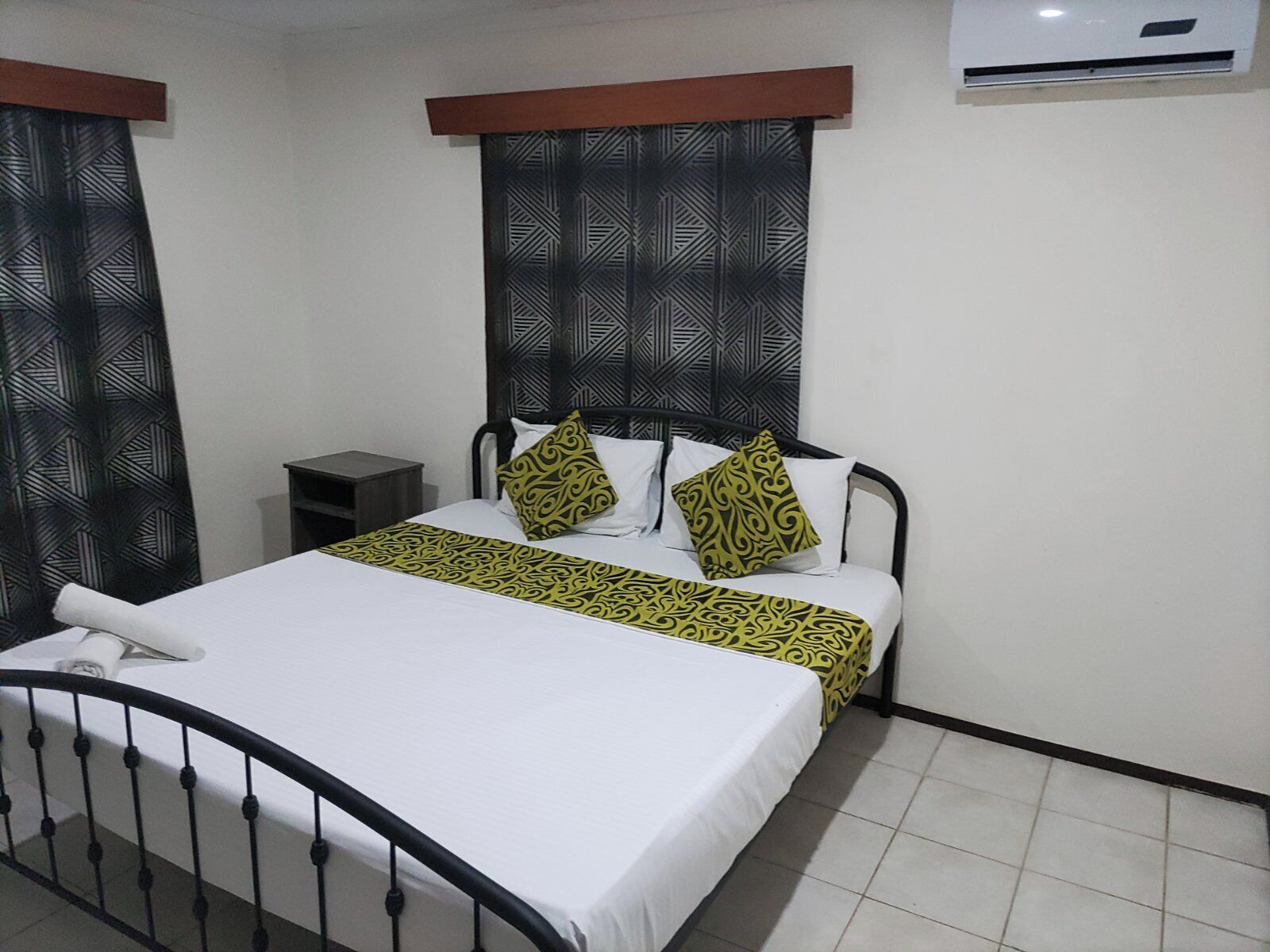 A single bed with a white mattress and a black metal frame. There is a small bedside table with a dark surface and a lamp. The room has a tiled floor and a window with a geometric patterned blind.