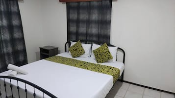 A single bed with a white mattress and a black metal frame. There is a small bedside table with a dark surface and a lamp. The room has a tiled floor and a window with a geometric patterned blind.