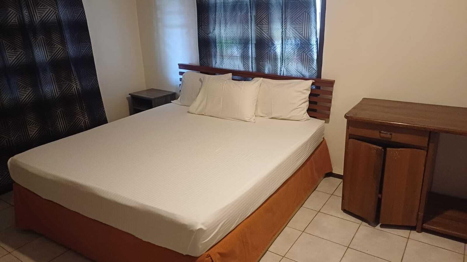 A bedroom with a bed, a nightstand, a wooden desk, and a window with curtains.