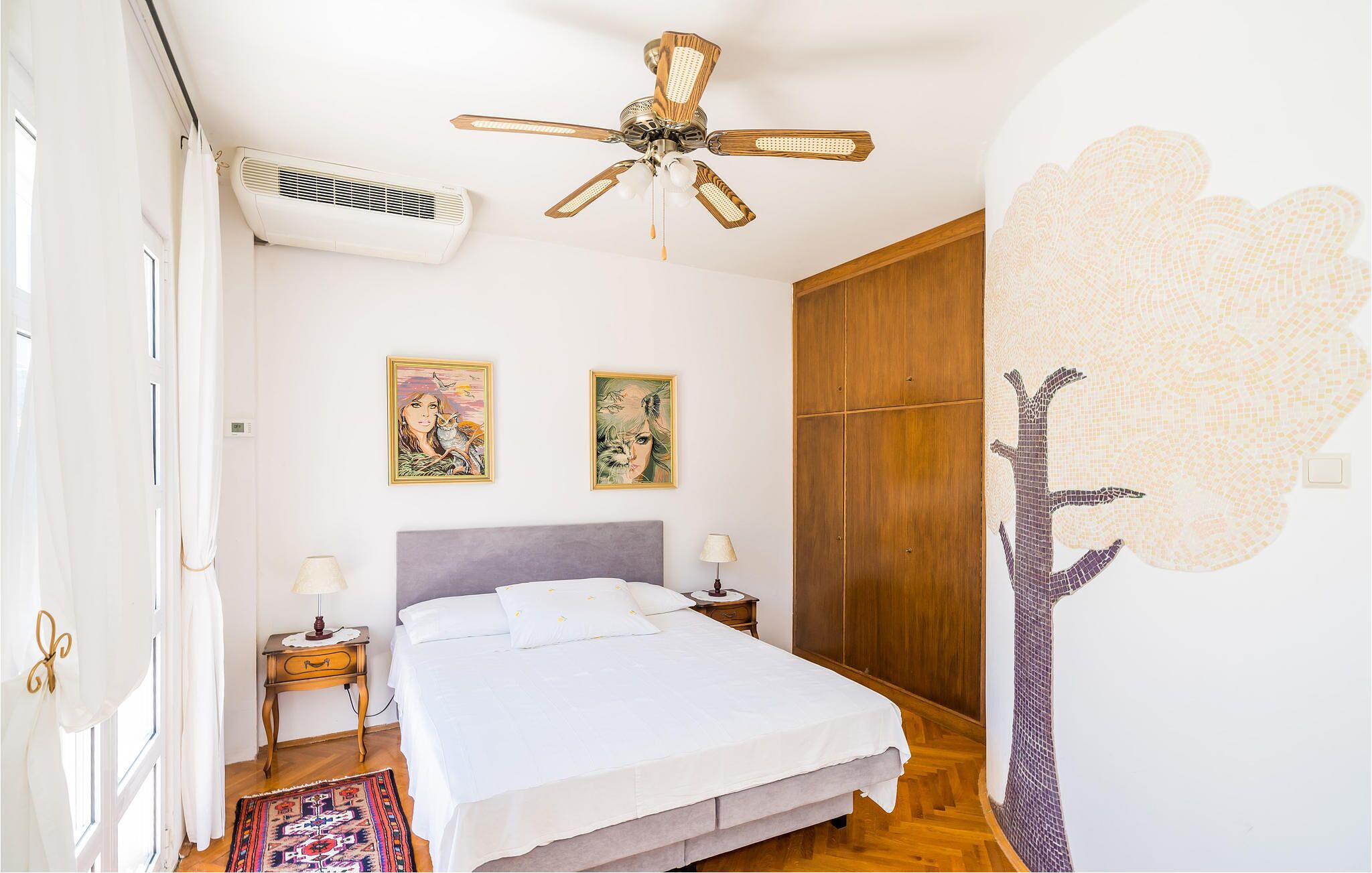 Un dormitorio con cama, dos aires acondicionados instalados en la pared, un ventilador de techo, una mesita de noche con lámpara, un armario de madera y dos obras de arte enmarcadas en la pared.