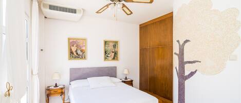 Un dormitorio con cama, dos aires acondicionados instalados en la pared, un ventilador de techo, una mesita de noche con lámpara, un armario de madera y dos obras de arte enmarcadas en la pared.
