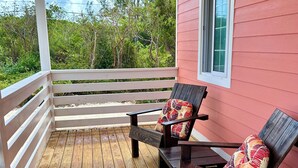 A wooden deck with two wooden chairs and a bench, a white railing, and a pink building with a window. - Mama Lou's Hide-A-Way   (Rainbow Bay)