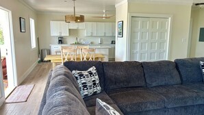 A modern living room with a grey sectional sofa, a wooden dining table with chairs, and a kitchen area with white cabinets and a ceiling fan. - Mama Lou's Hide-A-Way   (Rainbow Bay)