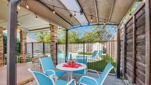 An outdoor patio area with a round table and blue chairs, a pool in the background, and string lights overhead. - 4 Bedroom & Pool: Great Irving Location close to attractions (Irving)